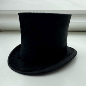 以下が含まれることがあります： 丸いブリムと高い円筒形のクラウンを備えた黒いトップハット。帽子のクラウンの基部には濃い色のバンドがあります。帽子はフェルトのような素材で作られています。