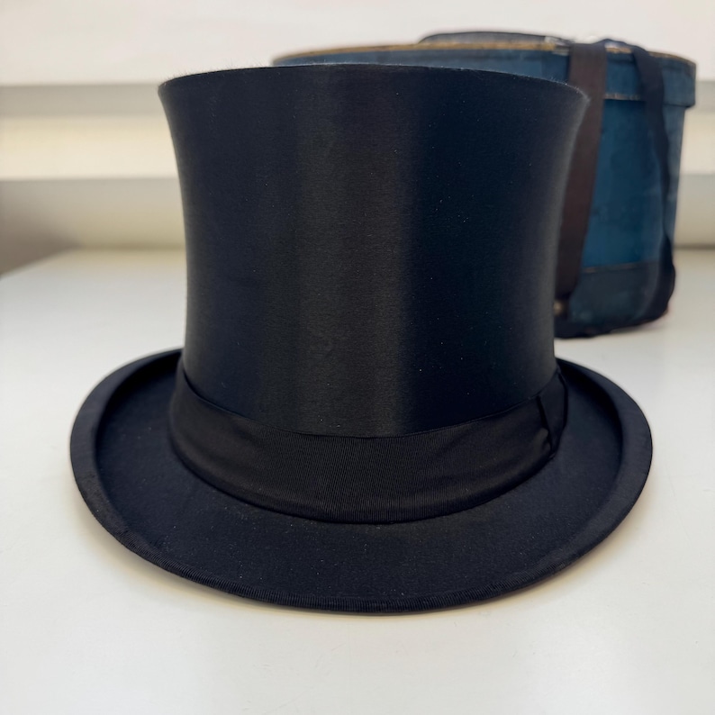 May include: A black top hat with a wide brim and a black band around the crown. The hat has a tall, cylindrical crown and a slightly curved brim. A blue box is in the background.