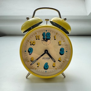 May include: A vintage yellow alarm clock with a cream-colored face. The clock features black hands, teal and yellow numerals, and the text "Youngline by Junghans". The clock has a curved handle and two yellow bells on top.