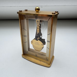 Puede incluir: Un reloj rectangular dorado con una esfera de cristal transparente. El reloj presenta números romanos y un mecanismo de péndulo visible en el interior. Una pequeña esfera dorada se encuentra en la parte superior.