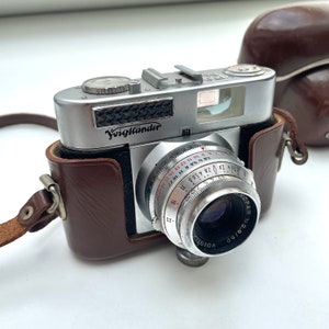 May include: A silver Voigtländer Vito B camera with a brown leather case and a strap. The camera has a lens with a black aperture and a silver lens cap. The camera has a silver dial with numbers and markings.