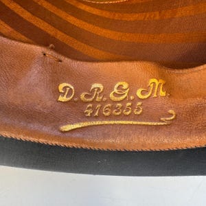 May include: Close-up of a hat's interior. The brown leather lining features gold lettering: "D.R. & M." and the number "416355". The hat's brim is black. The interior has a striped pattern.