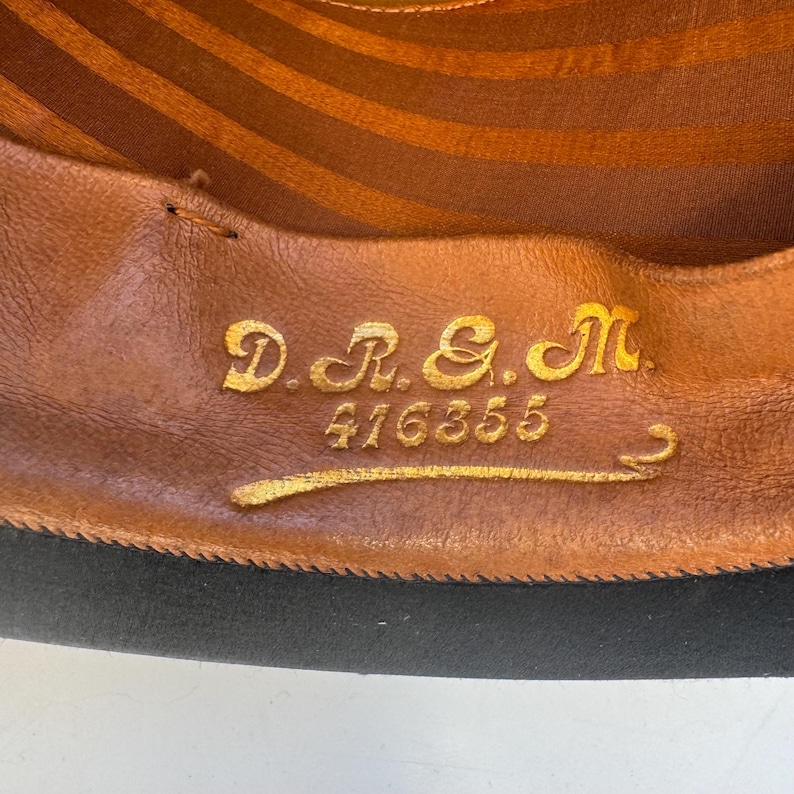 May include: Close-up of a hat's interior. The brown leather lining features gold lettering: "D.R. & M." and the number "416355". The hat's brim is black. The interior has a striped pattern.