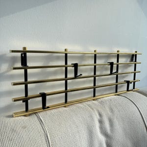 May include: A modern coat rack for wall mounting. It has horizontal brass bars held by vertical black metal supports. Black hooks are attached for hanging coats or other items. The rack is against a white wall.