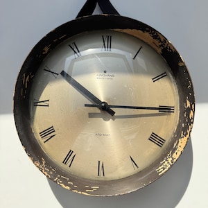 May include: Vintage Junghans electronic wall clock with a gold-toned face and black Roman numerals. The clock has black hands and a distressed, dark brown metal frame. The text "Junghans electronic" and "ATO-MAT" are visible on the face.