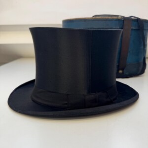 May include: A black top hat with a wide brim and a black band. The hat is on a white surface. A blue hat box is in the background.