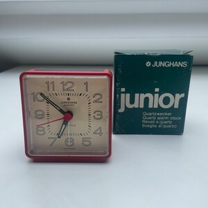 May include: A red alarm clock with a white face and black hands. The clock is labeled "JUNGHANS Junior" and has the text "Quartz alarm clock" on the box.