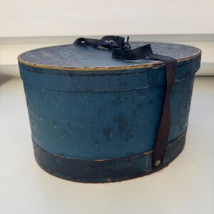 May include: A vintage blue hat box with a black ribbon and trim. The oval-shaped box has a distressed finish, with a gold accent on the rim. The box is made of cardboard or a similar material.