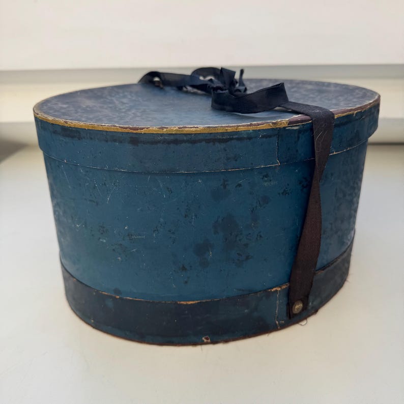 May include: A vintage blue hat box with a black ribbon and trim. The oval-shaped box has a distressed finish, with a gold accent on the rim. The box is made of cardboard or a similar material.