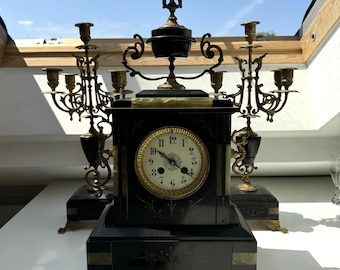 Könnte beinhalten: Eine schwarze Marmorkaminuhr mit goldenen Akzenten und einem römischen Zifferblatt. Die Uhr wird von zwei verzierten Kerzenleuchtern aus Messing mit schwarzen Akzenten flankiert.