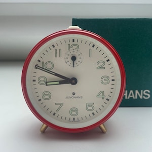 May include: A red alarm clock with a white face and black hands. The clock has a small second hand in the center and the words "JUNGHANS" and "MADE IN GERMANY" printed on the face.
