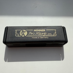 Könnte beinhalten: Ein schwarzes rechteckiges Etui mit goldfarbenen Buchstaben. Der Text auf dem Etui lautet "Hohner Pro Harp MS Made in Germany". Das Etui hat einen Klappdeckel und scheint aus Kunststoff zu sein. Das Etui ist für eine Mundharmonika.