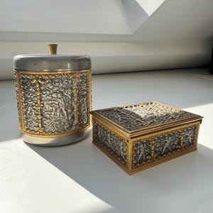 Op de afbeelding: Twee antieke decoratieve containers. Eén is een cilindervormige pot met een deksel en een kleine knop. De andere is een rechthoekige doos. Beide hebben zilver- en goudkleurige sierlijke bloemen- en figuurontwerpen.