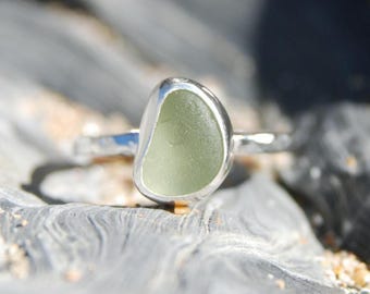 May include: A silver ring with a pale green sea glass stone. The stone is set in a polished silver band. The ring's design is simple and elegant. The sea glass has a smooth, frosted appearance. The ring is a beautiful piece of jewellery, suitable for any occasion.