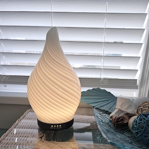 May include: A white teardrop-shaped essential oil diffuser with a spiraling ribbed texture. The base is black and features four small circular indicator lights with text labels reading 0.5H, 1H, 2H, and 3H printed in white on the front of the device.