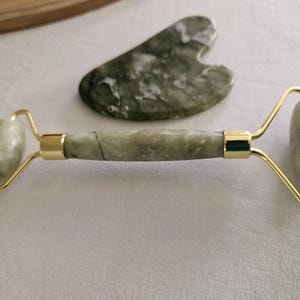 May include: A green jade facial roller with a gold handle and a matching green jade gua sha tool. The roller has two smooth, round jade stones that are used to massage the face and neck.