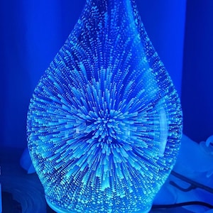 Könnte beinhalten: Blauer Glas-Diffusor für ätherische Öle in Tropfenform. Das Innere hat ein strukturiertes, sternförmiges Design, das von blauem Licht beleuchtet wird. Der schwarze Sockel hat vier kleine weiße Lichter.