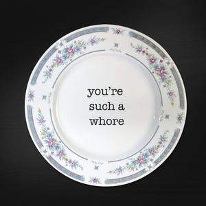 May include: A white ceramic dinner plate featuring a floral border pattern in shades of pink, blue, and green. The centre of the plate displays the black text 'you're such a whore' printed in a simple, clean, sans-serif typeface on the white surface.