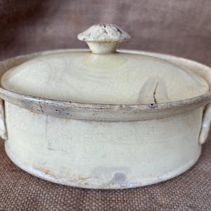 May include: Cream-coloured ceramic casserole dish with a lid. The dish is round with two handles and a lid with a knob. The rim and knob have a darker, aged appearance. The dish has a vintage, antique aesthetic.