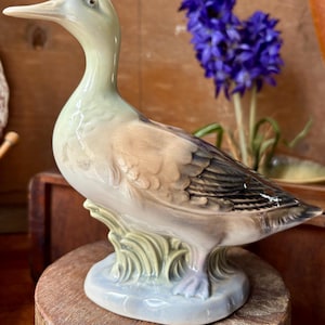 Könnte beinhalten: Keramik-Entenfigur mit langem Hals und detaillierten Federn. Die Ente hat einen hellgrünen Kopf und einen braunen Körper und steht auf einem hellblauen Sockel mit grünen Akzenten. Die Figur wird auf einer Holzoberfläche präsentiert.