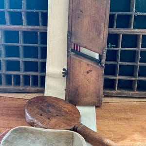 Könnte beinhalten: Eine Sammlung antiker Küchenutensilien, darunter eine Holzmandoline, ein rundes Holzwerkzeug mit Griff und eine Holzschaufel. Die Mandoline hat eine Klinge und einen rot-weiß gestreiften Akzent. Die Werkzeuge sind aus gealtertem Holz gefertigt.