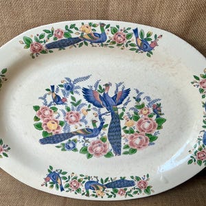 May include: An oval ceramic serving platter with a cream-coloured base featuring a central illustration of two blue peacocks surrounded by pink roses and green foliage, with additional peacocks and floral patterns along the outer rim.