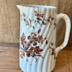 May include: A white ceramic jug with a vertical ribbed texture and a handle. The surface is decorated with a brown and purple floral pattern featuring leaves, blossoms, and a ribbon motif. The jug has a wide spout and a scalloped rim edge.