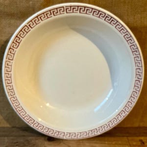 May include: A white ceramic bowl with a decorative border featuring a repeating Greek key pattern in a reddish-brown color. The bowl has a simple, classic design, suitable for serving various dishes.