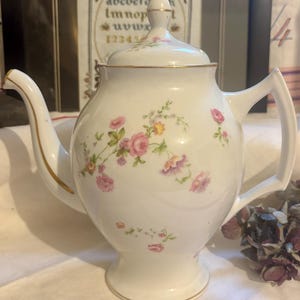 Può includere: Teiera in porcellana bianca con coperchio, ornata da motivi floreali rosa e viola e bordi dorati. La teiera presenta un beccuccio curvo e un manico. Un pezzo classico per servire il tè.