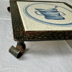 May include: A square trivet with a blue and white ceramic tile top featuring a decorative design. The tile is set in a dark metal frame with a distressed finish and curved legs. The trivet is designed to protect surfaces from heat.