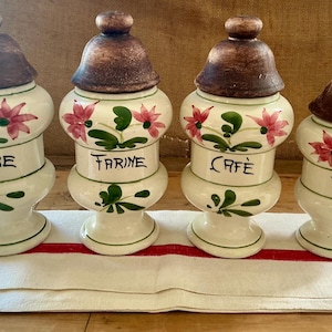 May include: Four ceramic kitchen canisters with brown lids and floral designs. The canisters are labeled "Sucre", "Farine", "Café", and "Poivre". The canisters are white with pink flowers and green leaves.