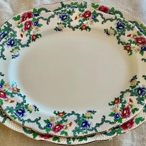 May include: Two oval ceramic platters featuring a white center and a decorative border with a green lattice pattern, accented by vibrant red, blue, and yellow floral motifs. The edges of the platters have a scalloped shape with a gold trim finish.