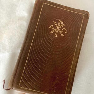 May include: A vintage brown leather-bound book with gold detailing. The cover displays a gold emblem within concentric circles. The book has a worn appearance, indicating age. A thin, light-coloured ribbon bookmark is visible.