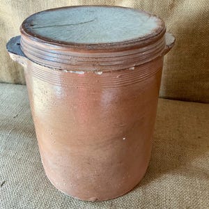 Può includere: Un vaso cilindrico in gres marrone con una finitura smaltata liscia e un coperchio abbinato con una parte superiore testurizzata di colore chiaro. Il contenitore presenta due piccole maniglie laterali e scanalature orizzontali sul bordo.