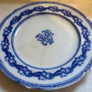 May include: White ceramic plate with a blue floral design. The plate has a scalloped edge and a detailed blue border with a repeating leaf pattern. A smaller floral design is in the center of the plate.