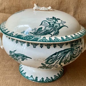 May include: A cream-colored ceramic tureen with a matching lid, featuring dark green floral patterns and decorative border designs. The tureen has two side handles, a pedestal base, and a sculpted handle on top of the domed lid.