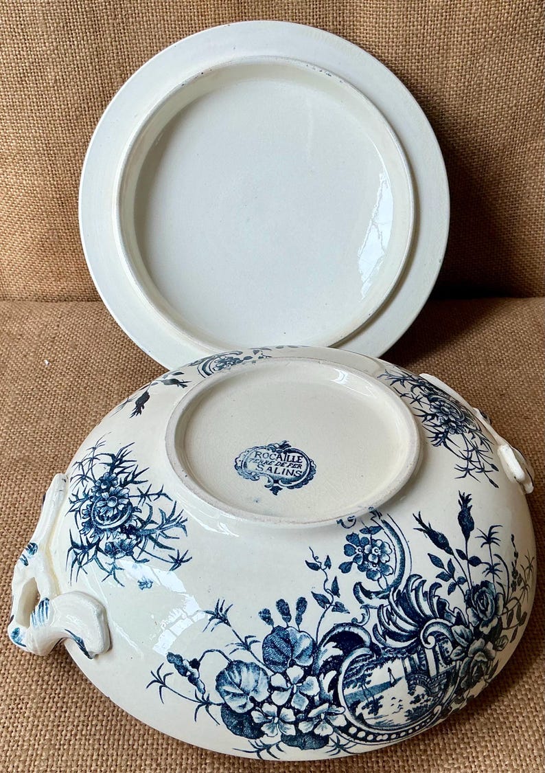 Puede incluir: Bol para servir de porcelana blanca con tapa, con un dise&ntilde;o floral azul. El bol tiene asas y un borde decorativo. La tapa es blanca lisa. El bol est&aacute; marcado con "Rocaille Terre de Fer, Salins".