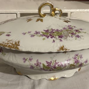 Puede incluir: Una fuente de porcelana blanca con tapa, adornada con motivos florales morados y detalles dorados. La fuente tiene un borde festoneado y ribete dorado. La tapa presenta un asa dorada. Una pieza clásica para servir alimentos.