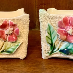 Op de afbeelding: Twee witte keramische bloempotten met reliëf roze bloemen en groene bladeren. De potten hebben een getextureerd oppervlak.