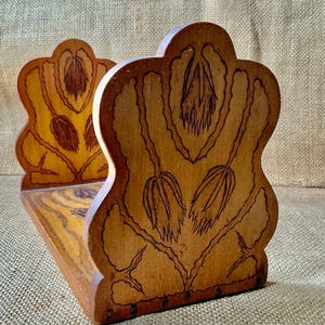 Puede incluir: Un sujetalibros de madera con acabado marrón claro y detalles de pirograbado en marrón oscuro. El diseño muestra un motivo floral con capullos alargados y caídos, además de tallos grabados en la superficie del panel de madera curvado.