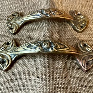 May include: Two antique brass drawer pulls with floral and leaf designs. Each pull has a curved handle with decorative details and mounting holes at each end. The brass has a warm, aged patina, suggesting vintage or antique hardware.