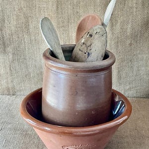 Może przedstawiać: Brązowy ceramiczny garnek umieszczony wewnątrz pasującej ceramicznej miski. Wewnątrz garnka znajdują się trzy drewniane łyżki o różnych odcieniach i fakturach. Na przedniej części miski znajduje się wytłoczone logo z napisem "Spong & Co".