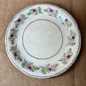 Op de afbeelding: Een witte keramische plaat met een bloemenontwerp. De plaat heeft een rand van kleine, donkerblauwe lijnen en een centrale ring van dezelfde kleur. Het bloemmotief bevat roze, gele en groene bloemen. De plaat heeft een licht verouderd uiterlijk.