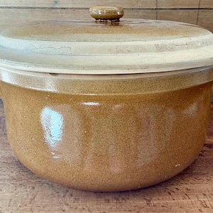 May include: A round, glazed ceramic casserole dish with a speckled tan and brown finish. The pot features a wide, flat, light-colored lid with a small, centered knob handle. The exterior of the vessel has a smooth, glossy texture throughout.