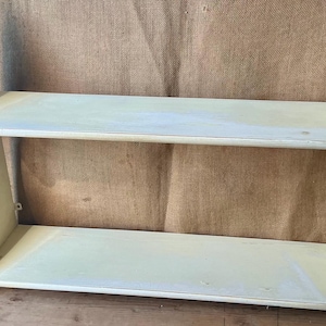 May include: A two-tiered, off-white wooden shelf with curved side supports. The shelves are rectangular and the overall design is simple and functional. The shelf is suitable for displaying items or for storage.