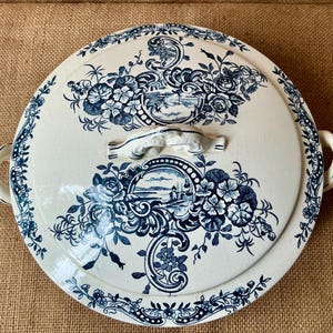 Puede incluir: Una fuente redonda de cer&aacute;mica blanca con tapa, adornada con motivos florales y paisaj&iacute;sticos azules. La fuente tiene dos asas y un asa central en la tapa. El borde y la tapa est&aacute;n decorados con intrincados dise&ntilde;os florales.