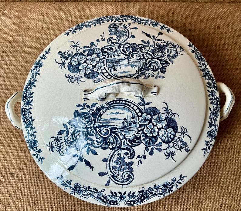 Puede incluir: Una fuente redonda de cer&aacute;mica blanca con tapa, adornada con motivos florales y paisaj&iacute;sticos azules. La fuente tiene dos asas y un asa central en la tapa. El borde y la tapa est&aacute;n decorados con intrincados dise&ntilde;os florales.