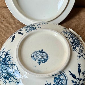 Puede incluir: Fuente de cer&aacute;mica color crema con tapa, con un estampado floral azul. La fuente tiene forma redondeada con un borde ligeramente elevado y asas. La parte inferior de la fuente tiene un sello azul que dice "Rocaille Terre de Fer Salins."