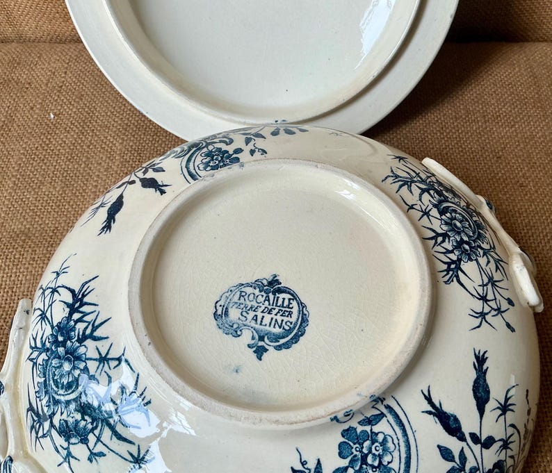 Puede incluir: Fuente de cer&aacute;mica color crema con tapa, con un estampado floral azul. La fuente tiene forma redondeada con un borde ligeramente elevado y asas. La parte inferior de la fuente tiene un sello azul que dice "Rocaille Terre de Fer Salins."