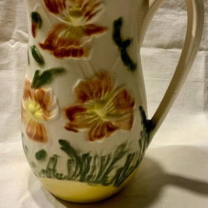 May include: A vintage ceramic pitcher with a floral design. The pitcher is white with brown, yellow, and green flowers. The pitcher has a wide mouth and a handle.